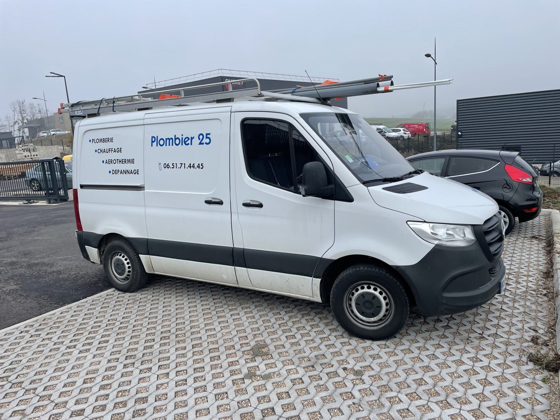 Fourgonnette de service avec échelle sur le toit, garée sur un parking goudronné à côté d'une voiture.