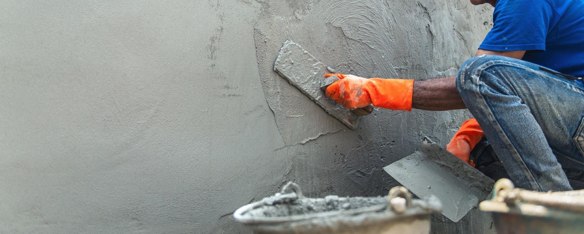 Ein Arbeiter in orangefarbenen Handschuhen glättet mit einer Kelle nassen Beton an einer Wand.