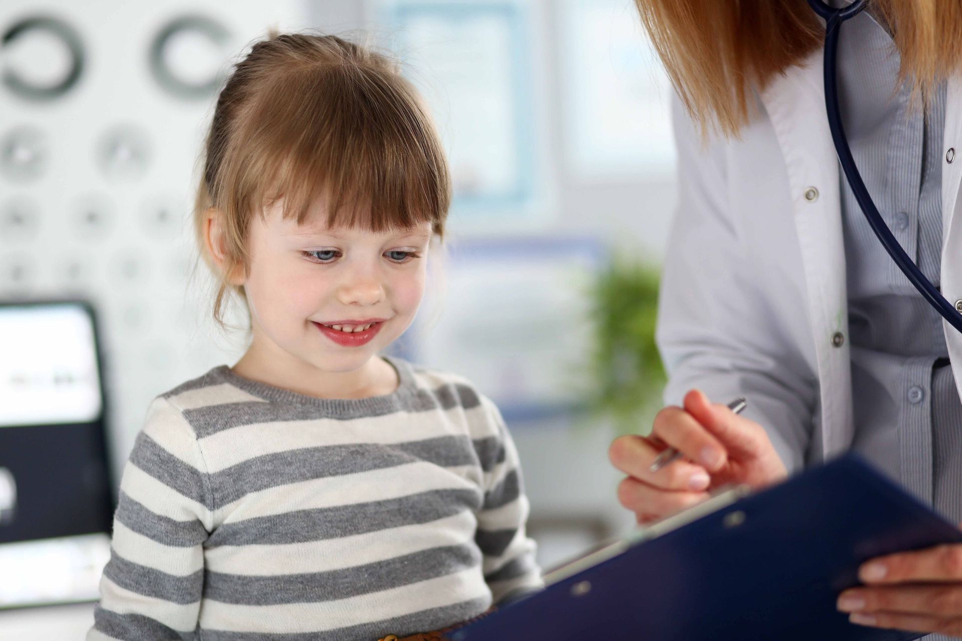 Ein kleines Mädchen sitzt neben einem Arzt und schaut auf ein Klemmbrett.