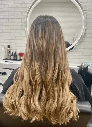 Una mujer con cabello largo y rubio está sentada en una silla frente a un espejo.