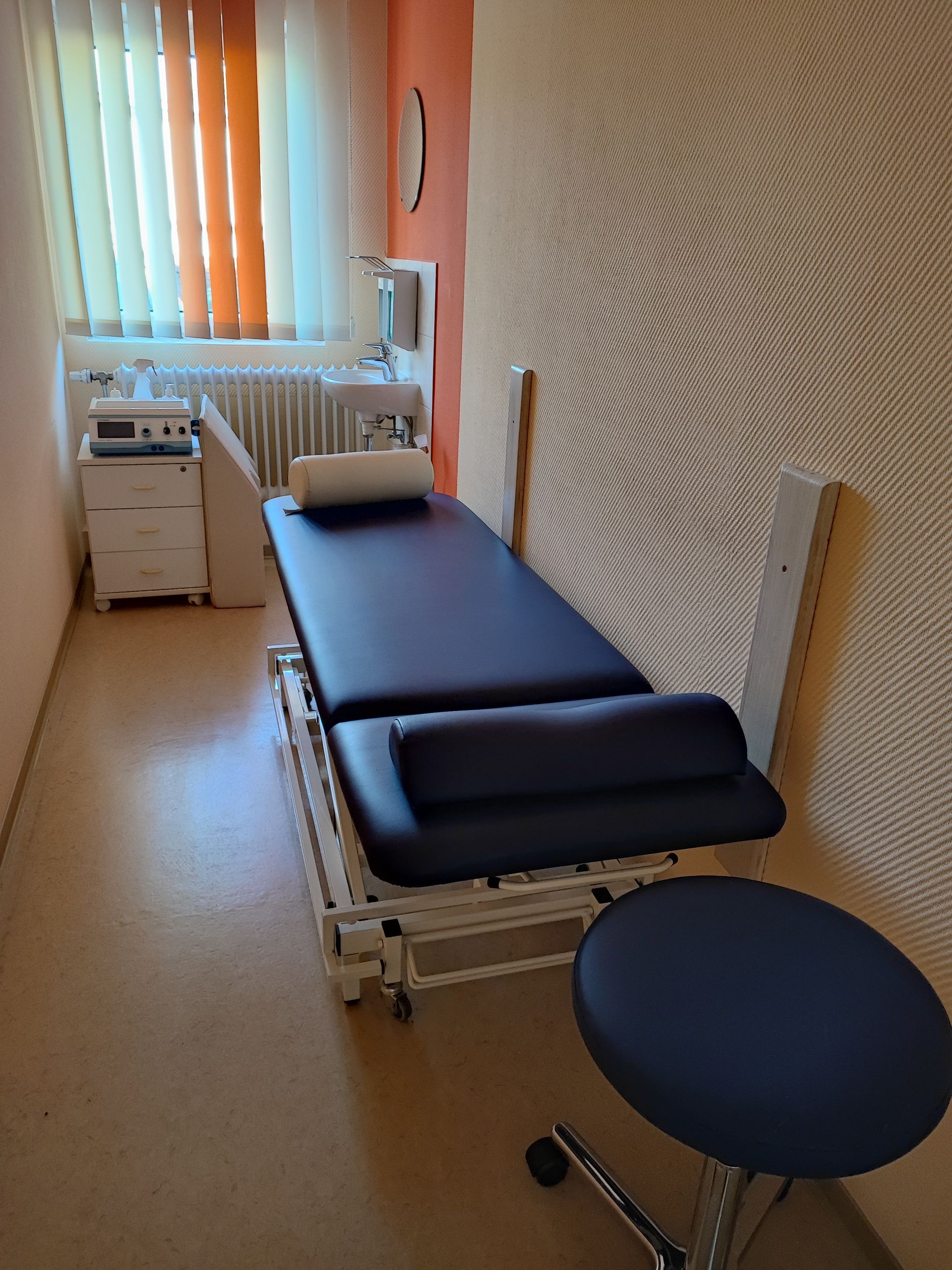 Untersuchungsraum mit Massageliege, Hocker und medizinischen Geräten.
