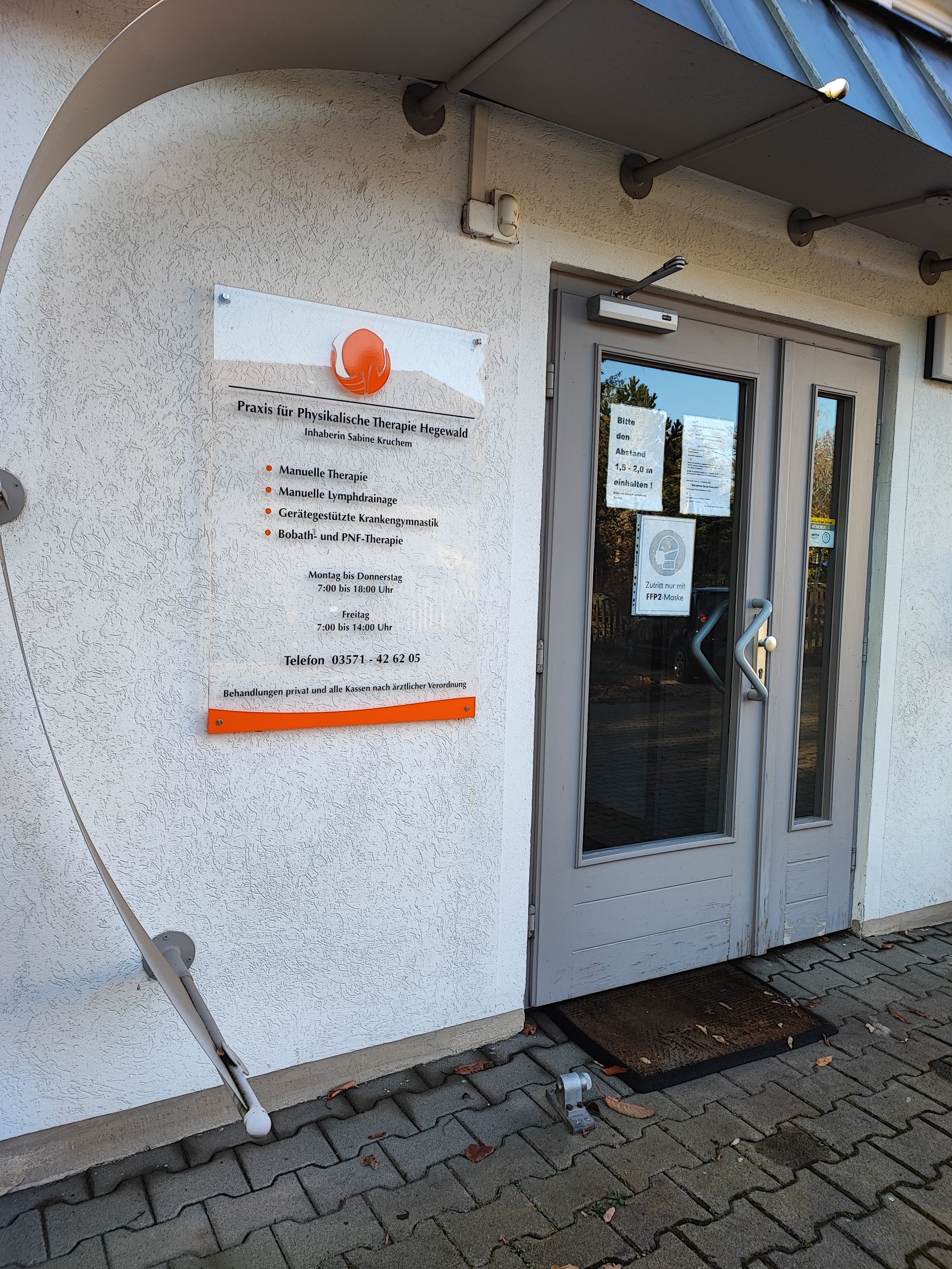 Außenansicht eines Gebäudes mit Glastür und Beschilderung. Über dem Text auf einer transparenten Platte befindet sich ein orangefarbenes Logo.