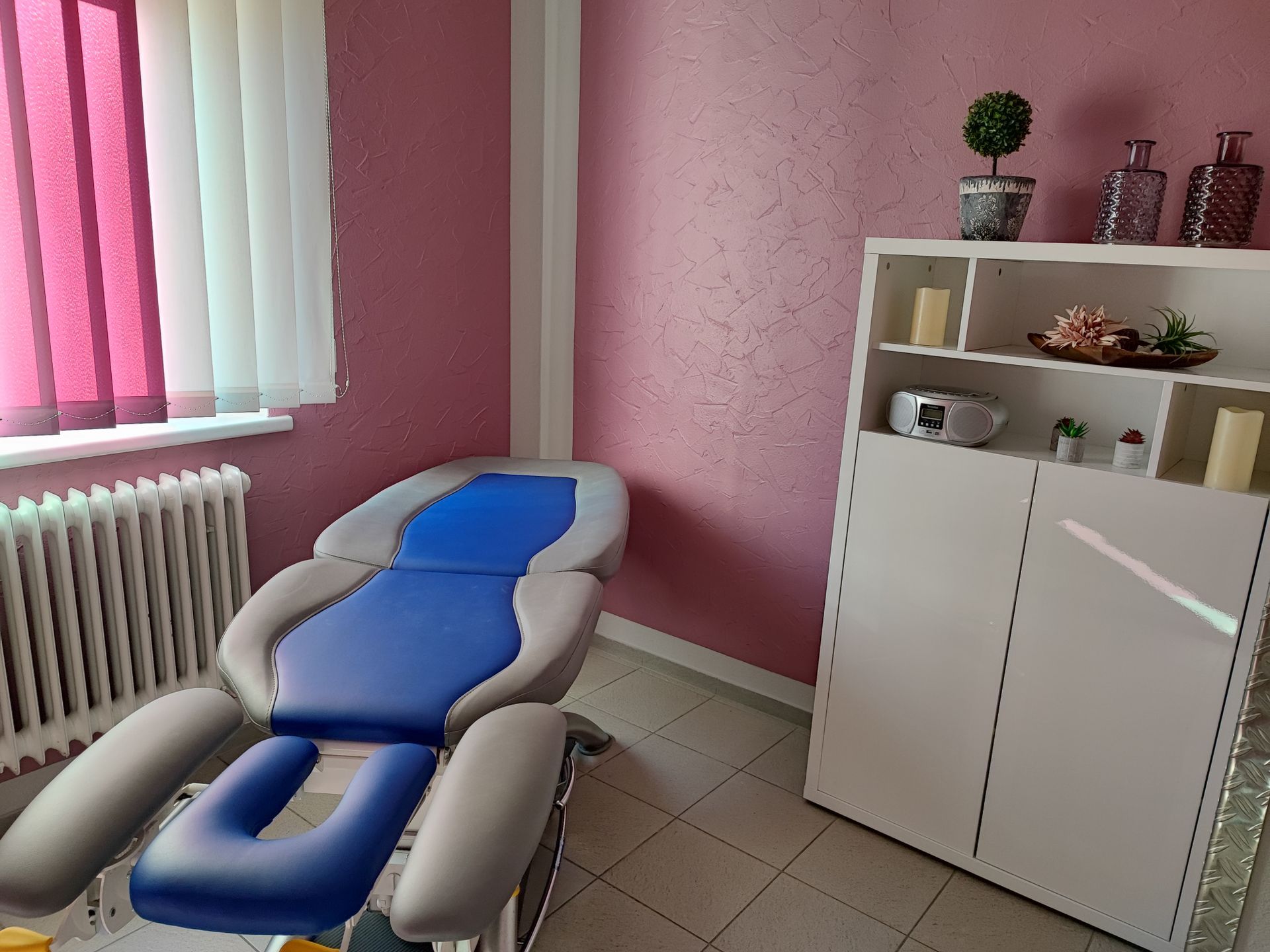 Behandlungsraum mit einem blau-grauen Stuhl, einem weißen Schrank, einem Fenster und rosa Wänden.