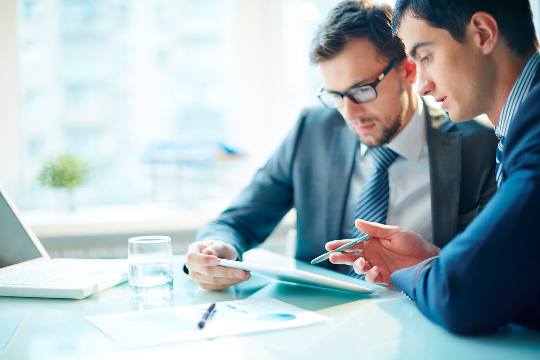 Dos hombres de traje revisando documentos en un escritorio, uno apuntando a una tableta, luz solar.