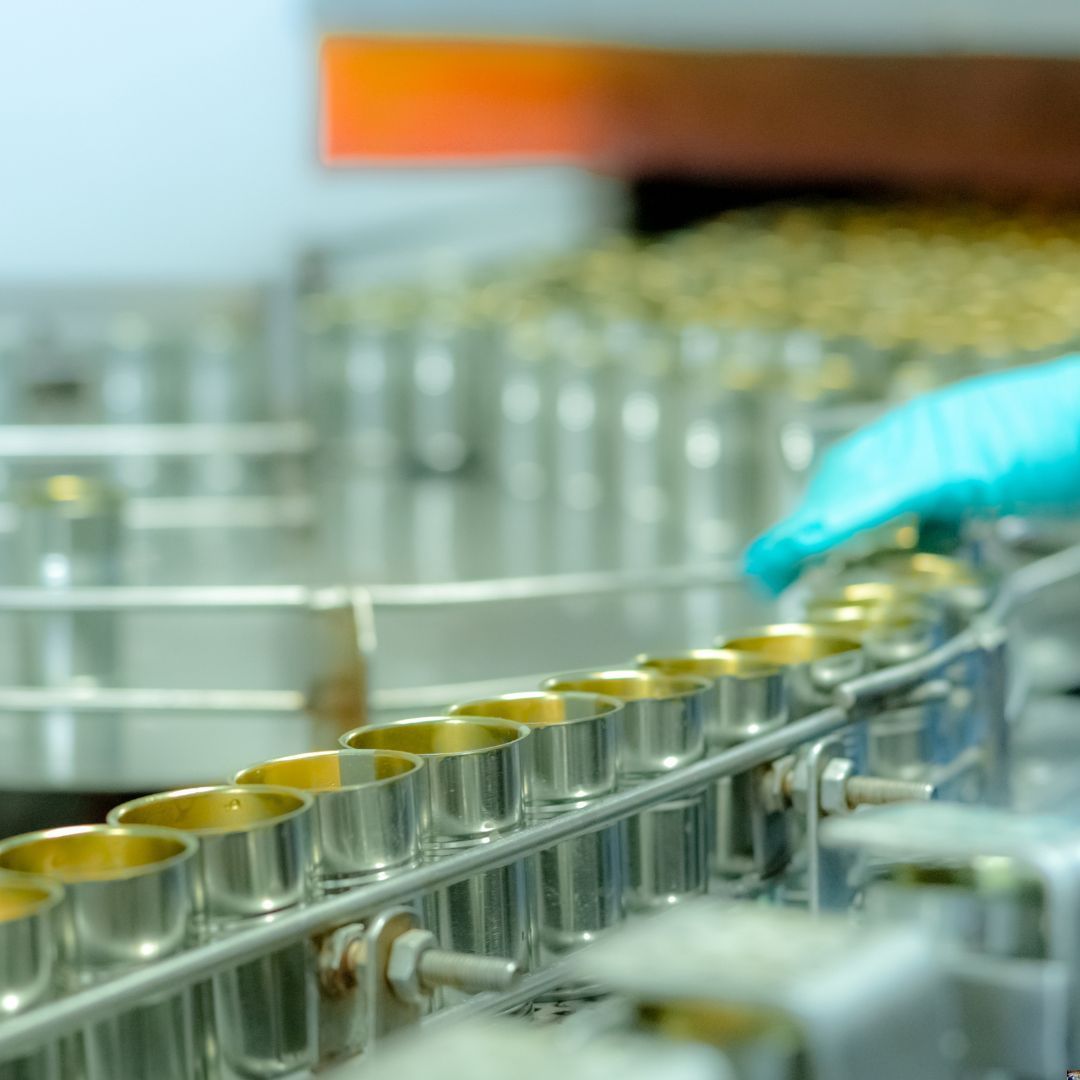 Latas de metal para alimentos que se llenan en una línea de montaje de una fábrica, con una mano enguantada revisándolas.