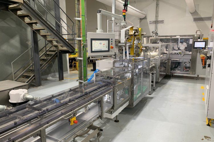 Maquina envasadora industrial en entorno de fábrica; acero inoxidable, cinta transportadora, escaleras.