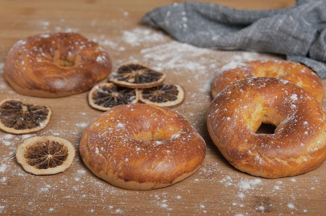 Bagels dorados espolvoreados con harina sobre una superficie de madera con rodajas de cítricos secos y un paño.
