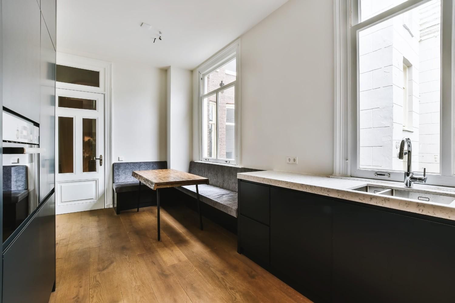 Cocina luminosa y minimalista con suelo de madera, armarios negros y grandes ventanales con zona de mesa central.