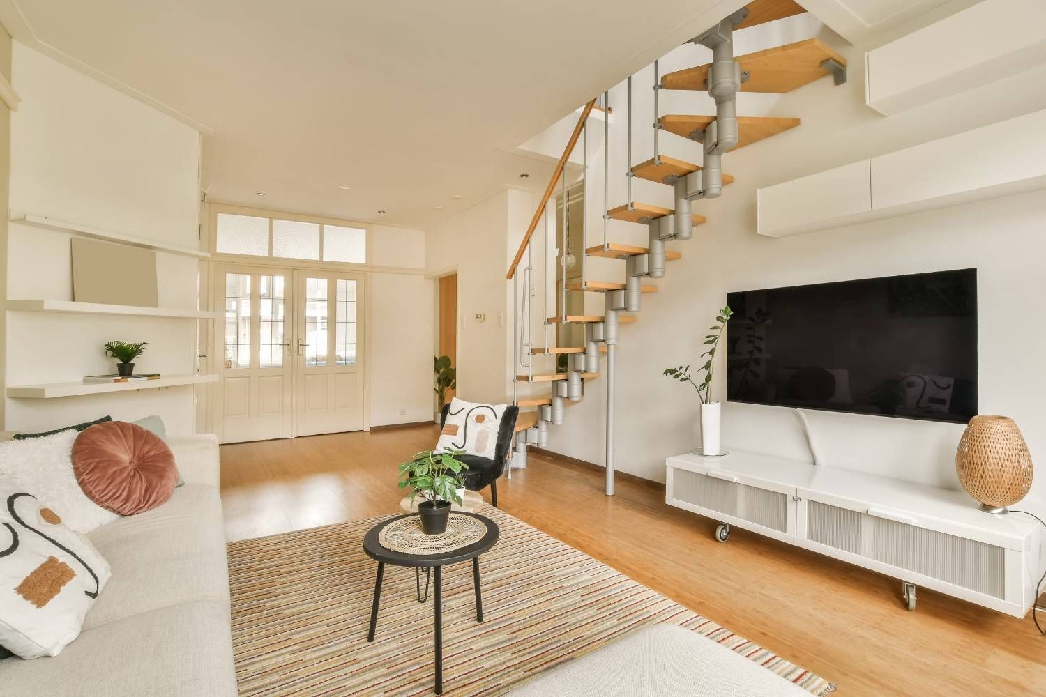 Luminoso salón con sofá blanco, escalera, mueble para la televisión y alfombra tejida en tonos cálidos de madera.