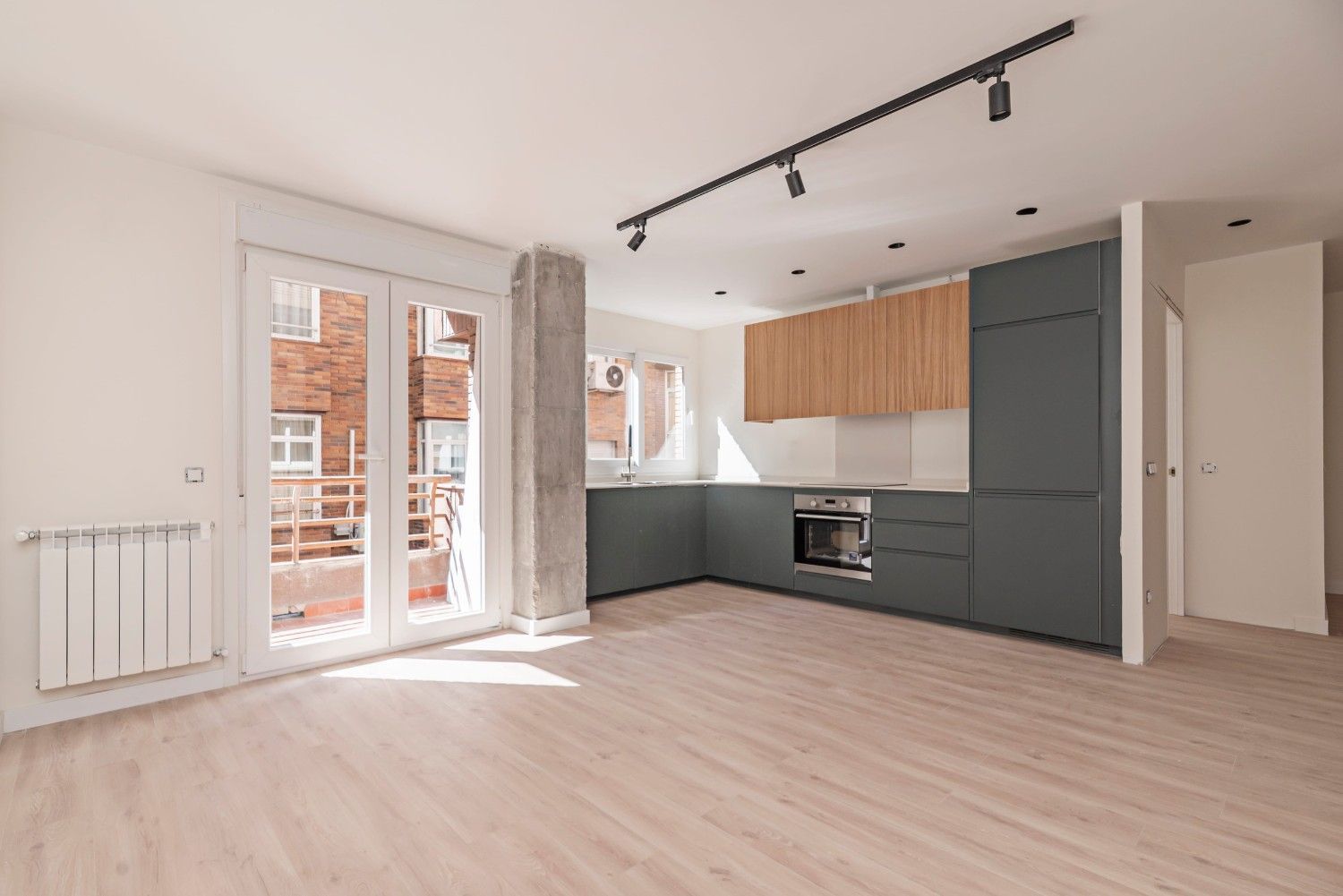 Apartamento moderno, luminoso y vacío, con suelos de madera clara, cocina gris y puertas de cristal que dan a un balcón.
