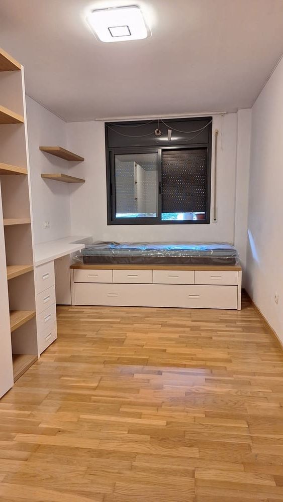 Dormitorio pequeño y luminoso con cama empotrada, escritorio, estanterías y ventana con persiana negra; suelo de madera.