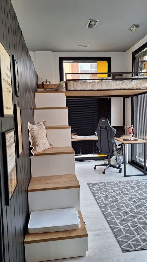 Cama alta en una habitación moderna. Escaleras que suben a la cama. Escritorio y silla debajo. Alfombra gris y pared negra con acentos.