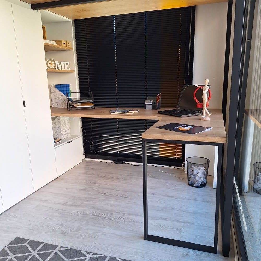 Escritorio en forma de L en una oficina en casa con una computadora portátil, almacenamiento y persianas; tonos grises y madera.