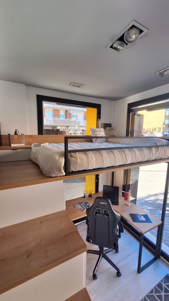 Cama alta con escritorio, escalera y ventana grande en una habitación pequeña y moderna. Combinación de colores madera clara, blanco y negro.