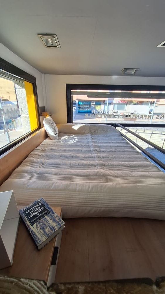 Dormitorio en una furgoneta camper con una cama junto a una ventana y un libro en una mesita de noche.