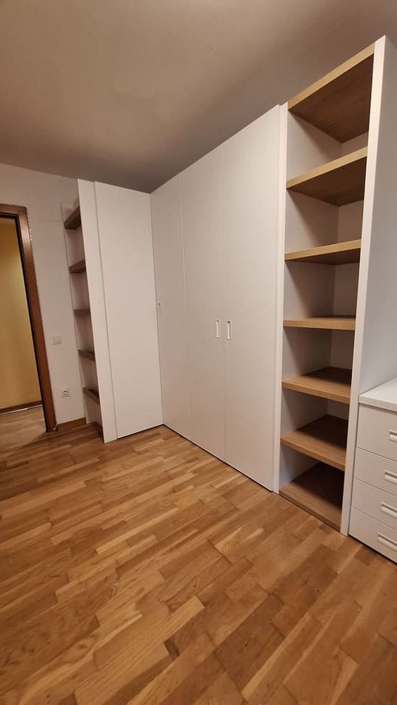 Armario empotrado blanco con baldas abiertas de madera y puertas cerradas, ubicado en una habitación con suelo de madera.