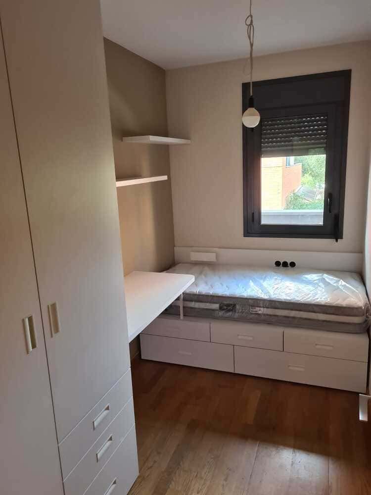 Dormitorio pequeño con muebles blancos: cama con cajones, escritorio, armario y estantes; piso de madera marrón; pequeña ventana.