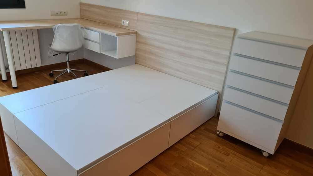 Dormitorio con cama blanca, escritorio, silla y cómoda. Cabecero con vetas de madera y suelo de madera.
