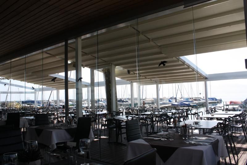 Le restaurant avec tables et chaises sous une verrière avec des bateaux en arrière-plan.