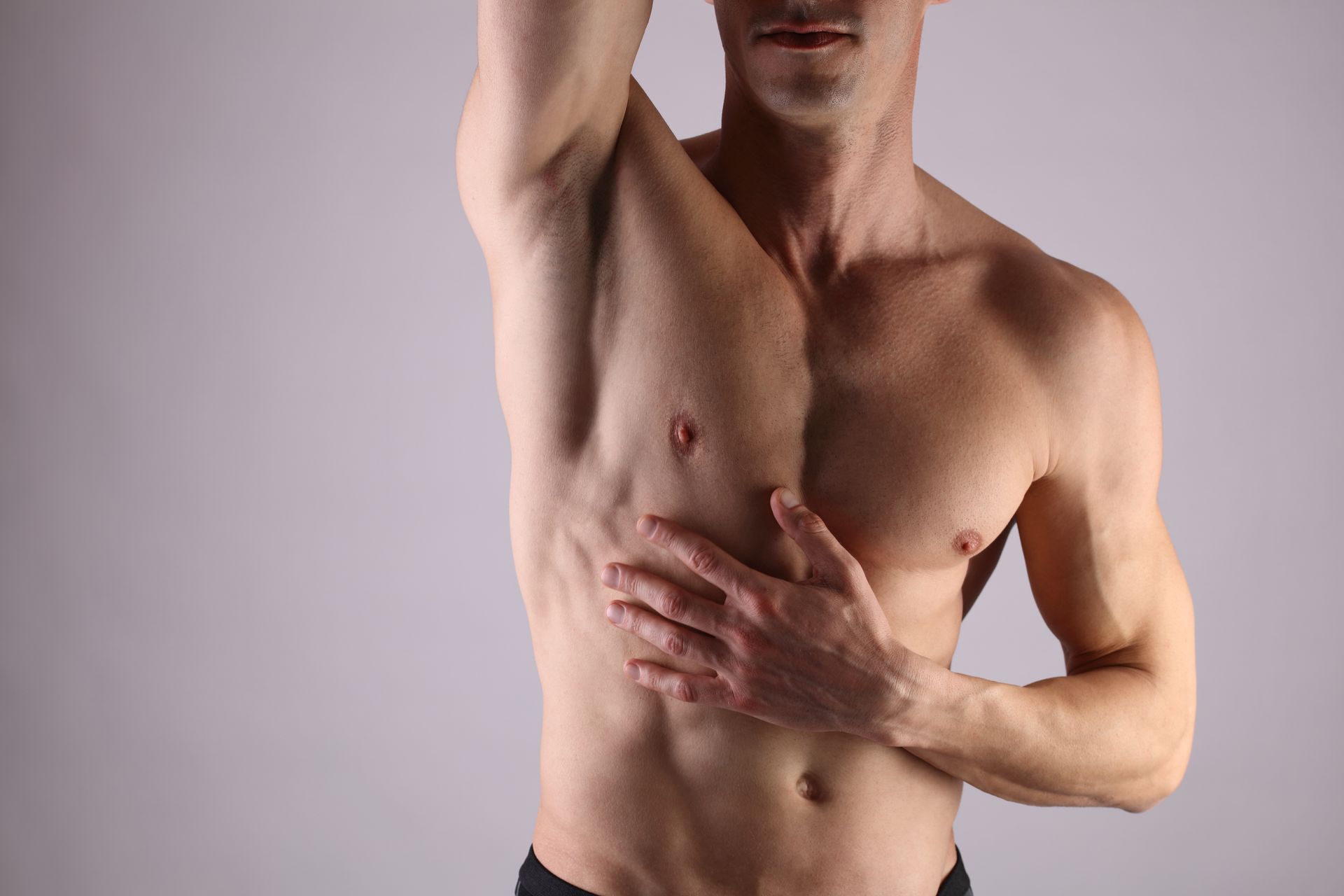 Homme musclé torse nu posant avec un bras levé sur fond gris uni.