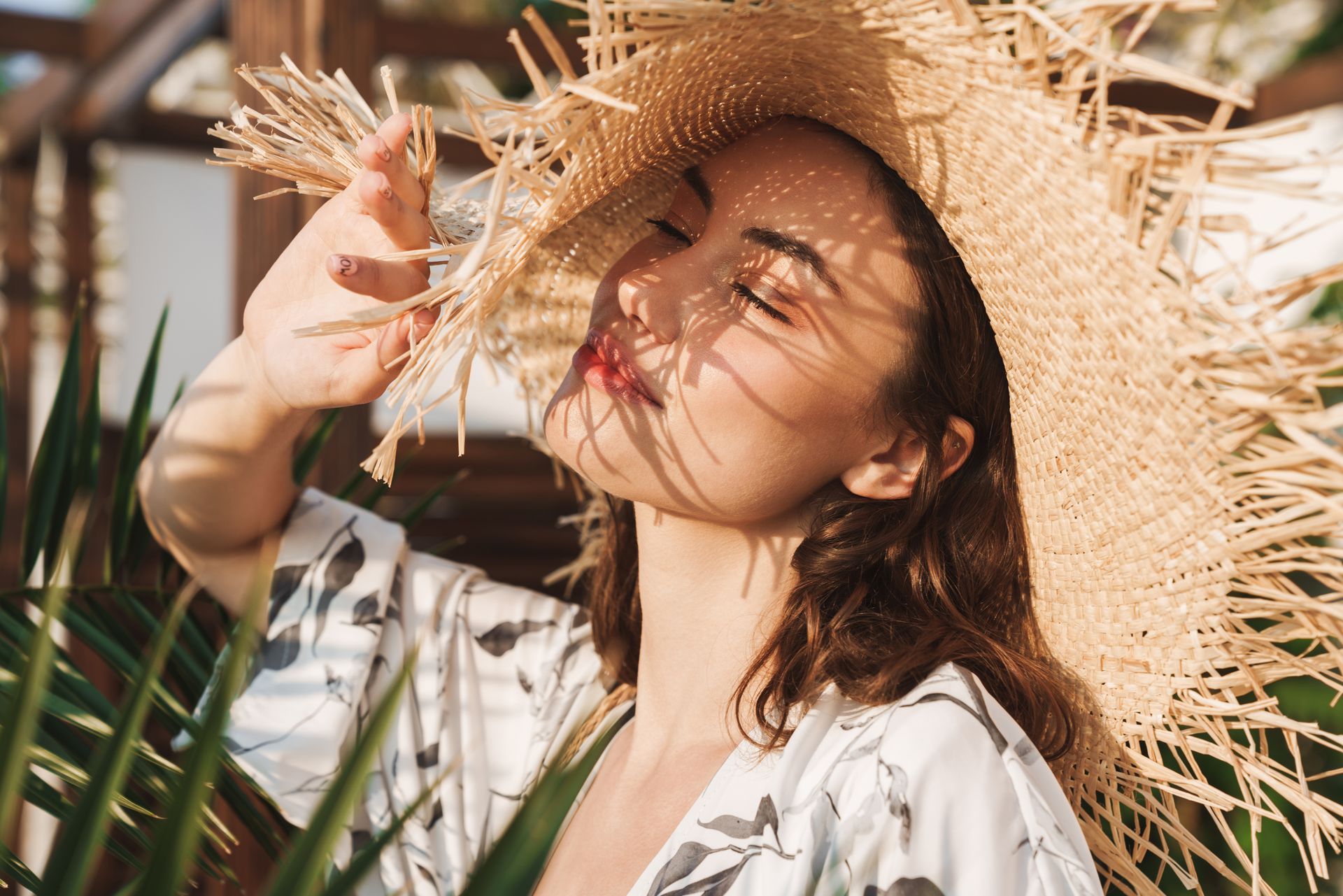 Femme coiffée d'un chapeau de paille dont elle tient le bord sous un soleil radieux, avec des feuilles de palmier en arrière-plan.