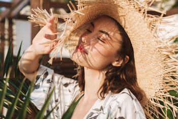 Femme coiffée d'un chapeau de paille dont elle tient le bord sous un soleil radieux, avec des feuilles de palmier en arrière-plan.