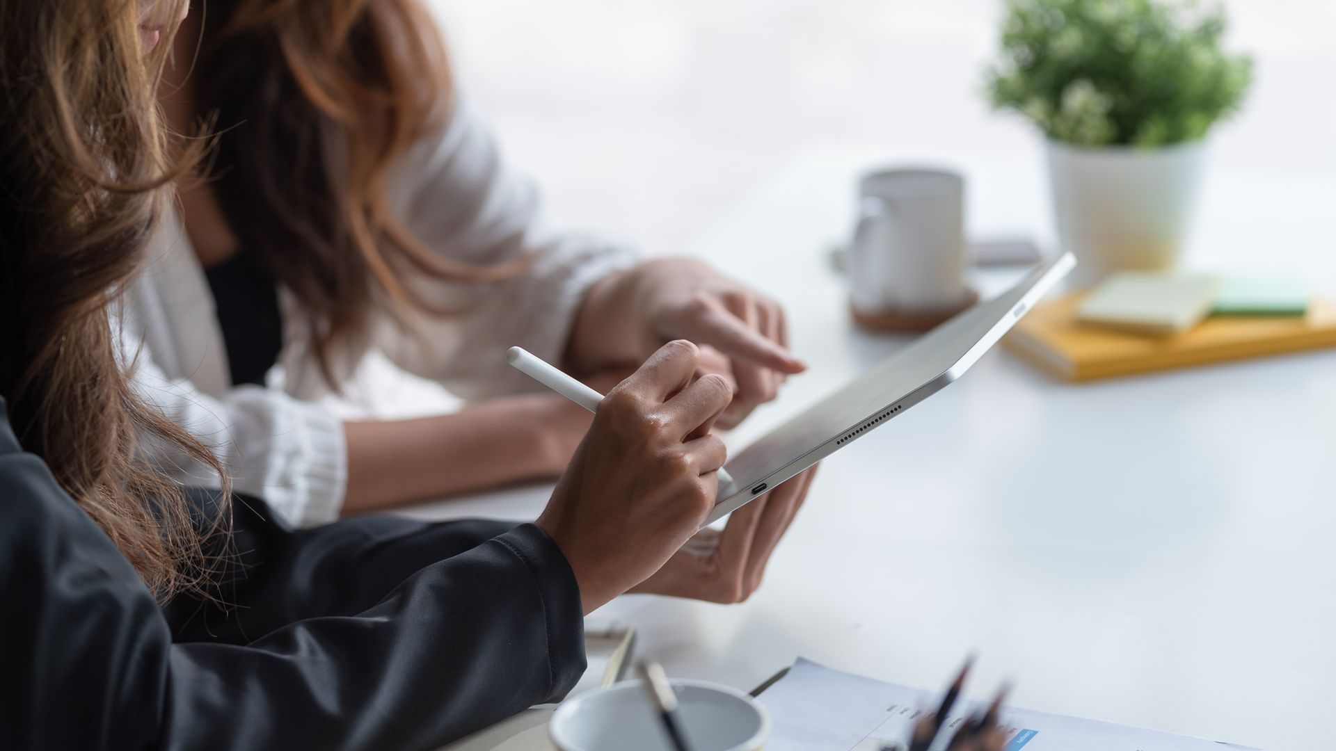 Une personne écrit sur une tablette à un bureau, avec une tasse de café et une petite plante en pot à proximité.