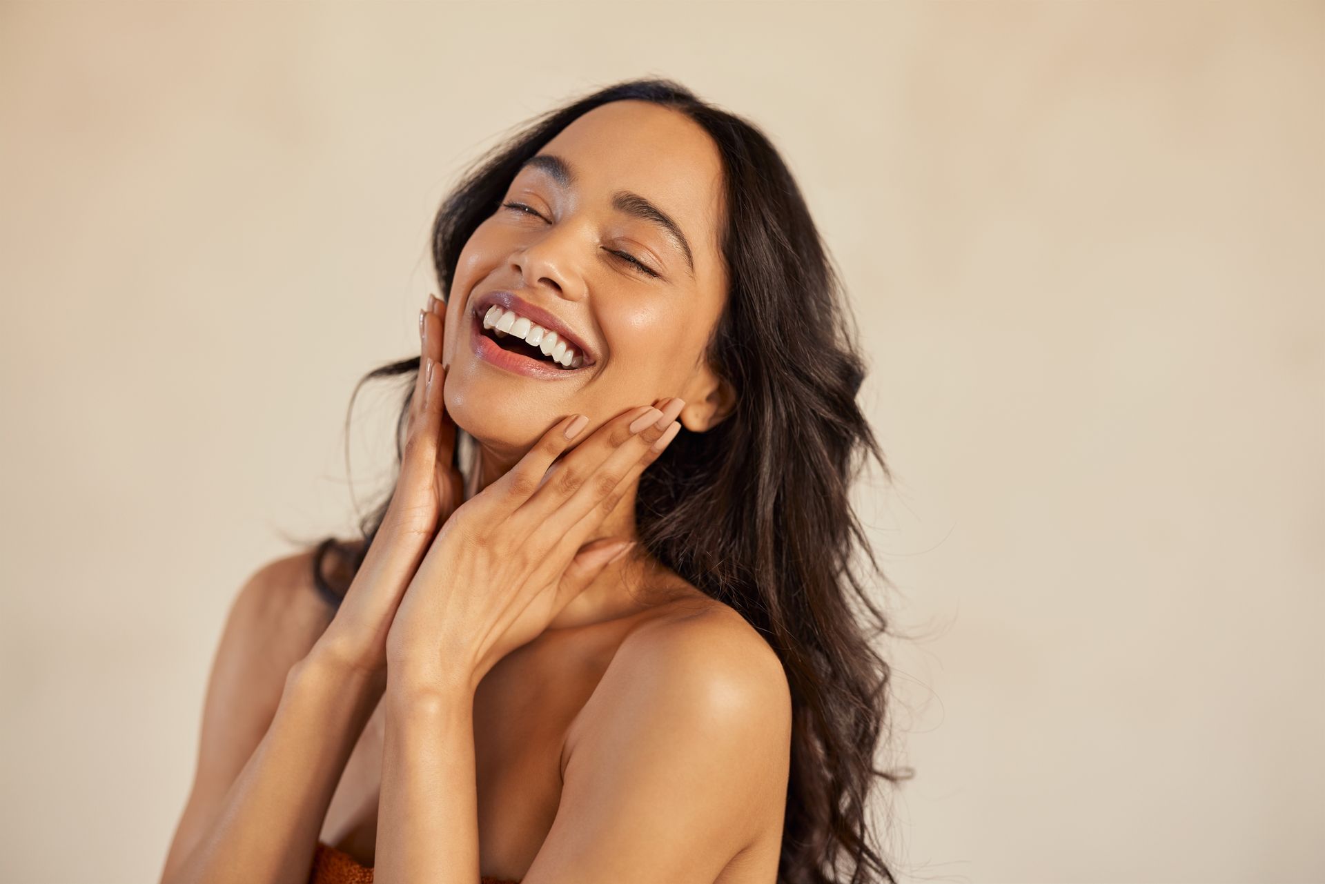 Personne souriante aux longs cheveux noirs touchant sa nuque sur fond beige.