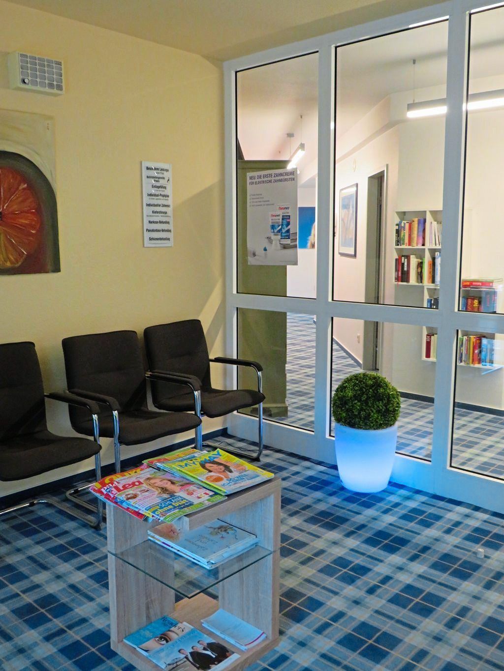 Der helle Eingangsbereich oder Flur der Praxis mit Blick durch eine Glastür auf ein Bücherregal und Hinweisschilder.