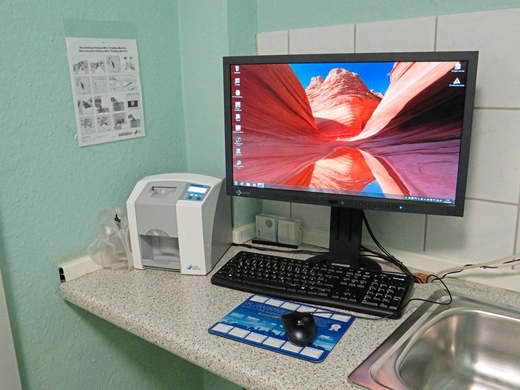 Ein Computerarbeitsplatz in der Praxis mit Monitor, Tastatur und einem medizinischen Gerät auf einer hellen Arbeitsplatte.