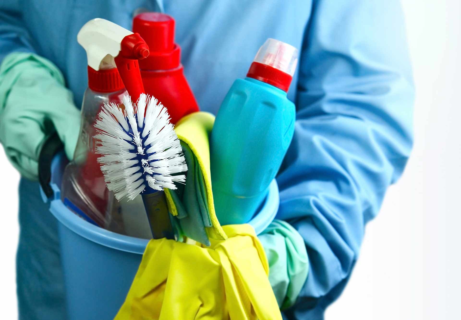 Des mains gantées tenant un seau de produits de nettoyage colorés et des brosses