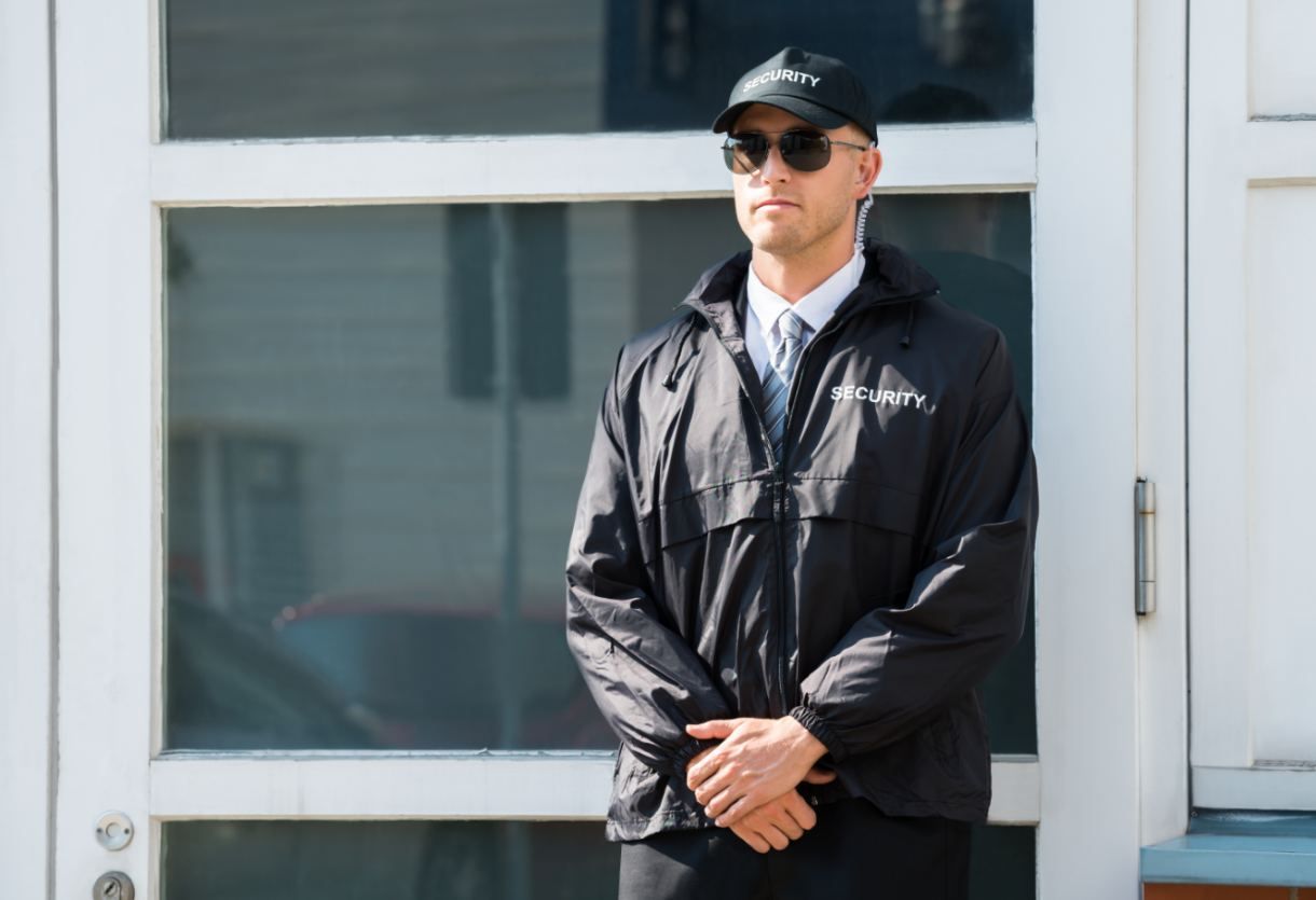 Un homme en veste et casquette noires se tient près de la fenêtre d'un immeuble, les mains jointes, portant des lunettes de soleil.