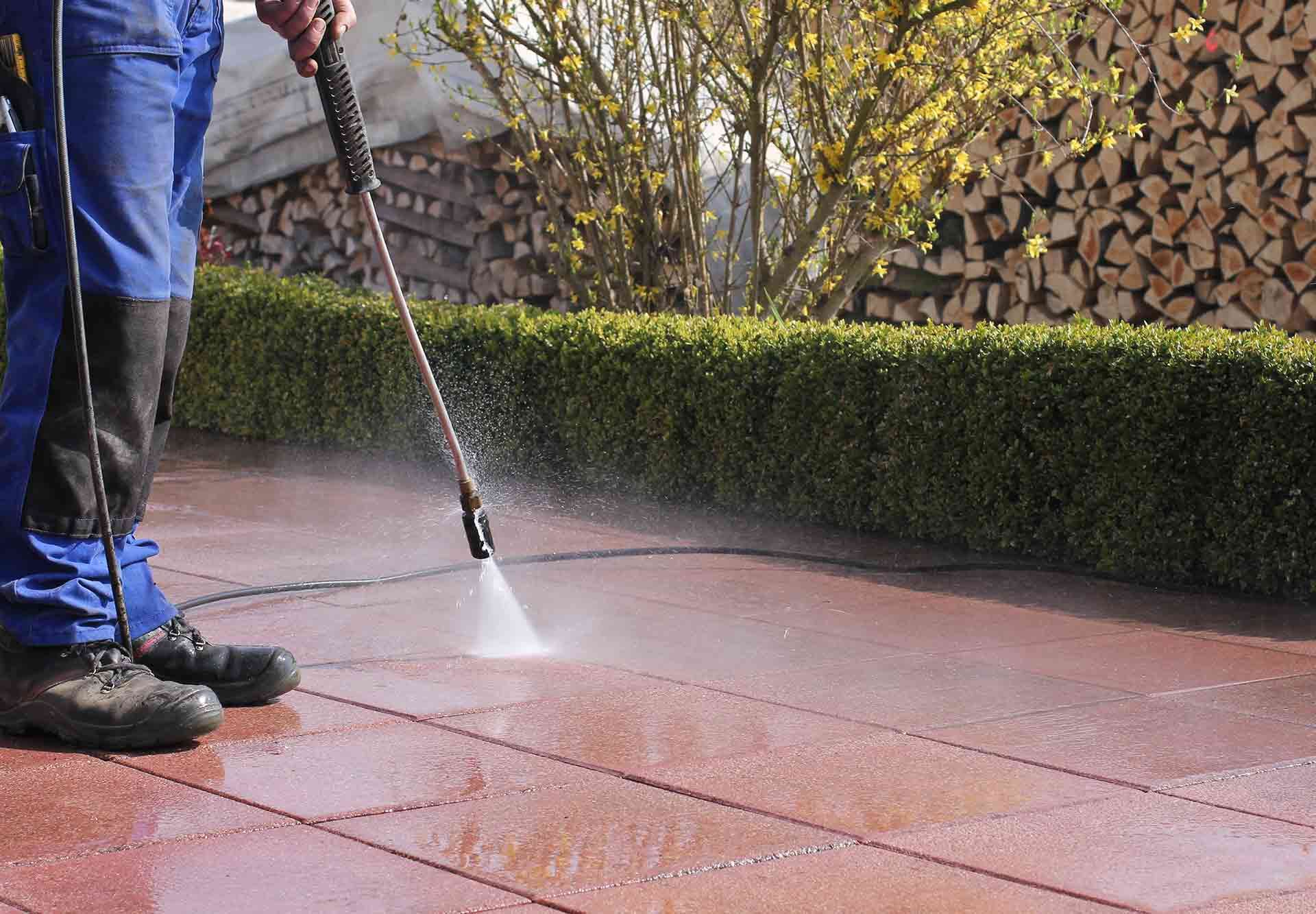 Une personne nettoyant au nettoyeur haute pression une terrasse rouge à côté d'une haie verte et de bois de chauffage empilé.