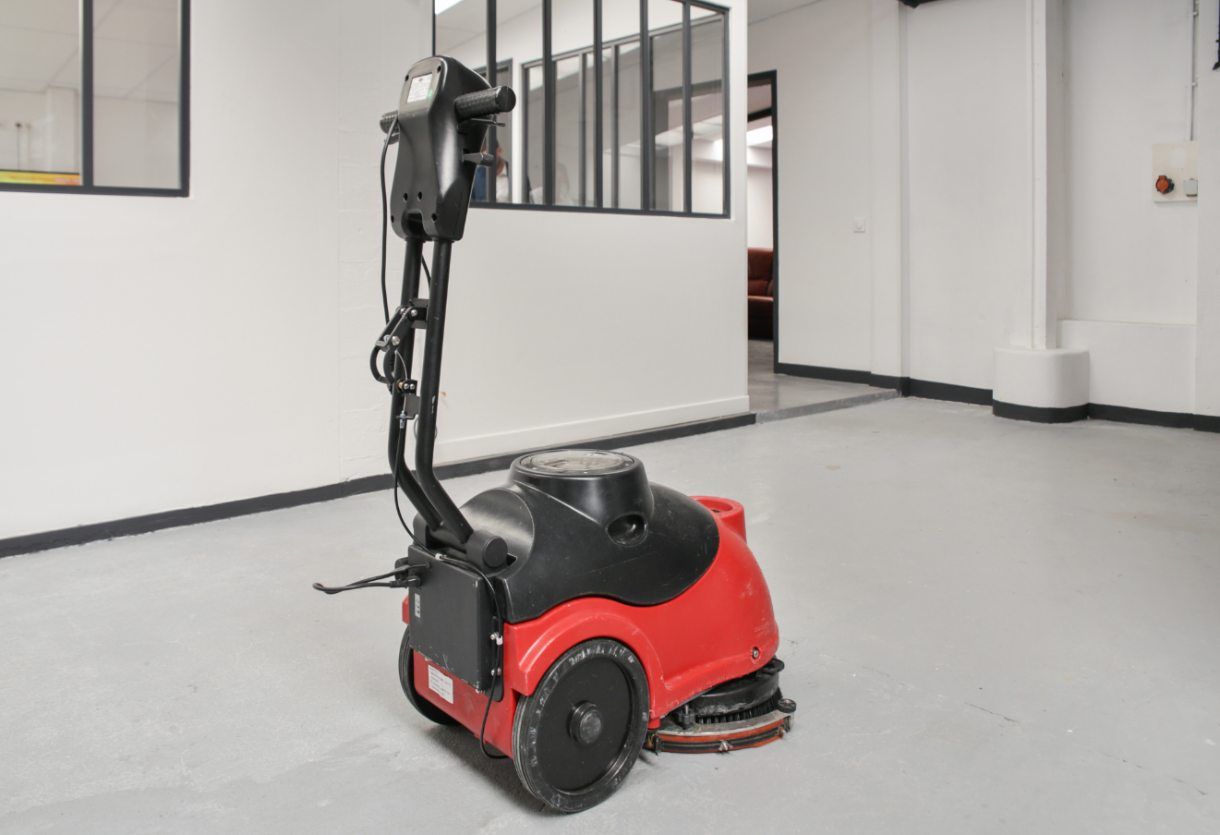 Machine de nettoyage de sol rouge dans un couloir blanc avec des cloisons de bureau en verre.