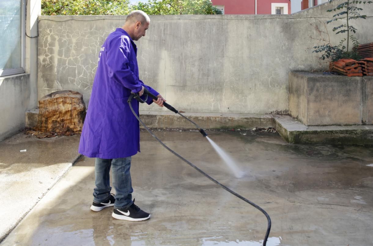 Une personne en manteau violet nettoie à haute pression une terrasse en béton à l'extérieur.