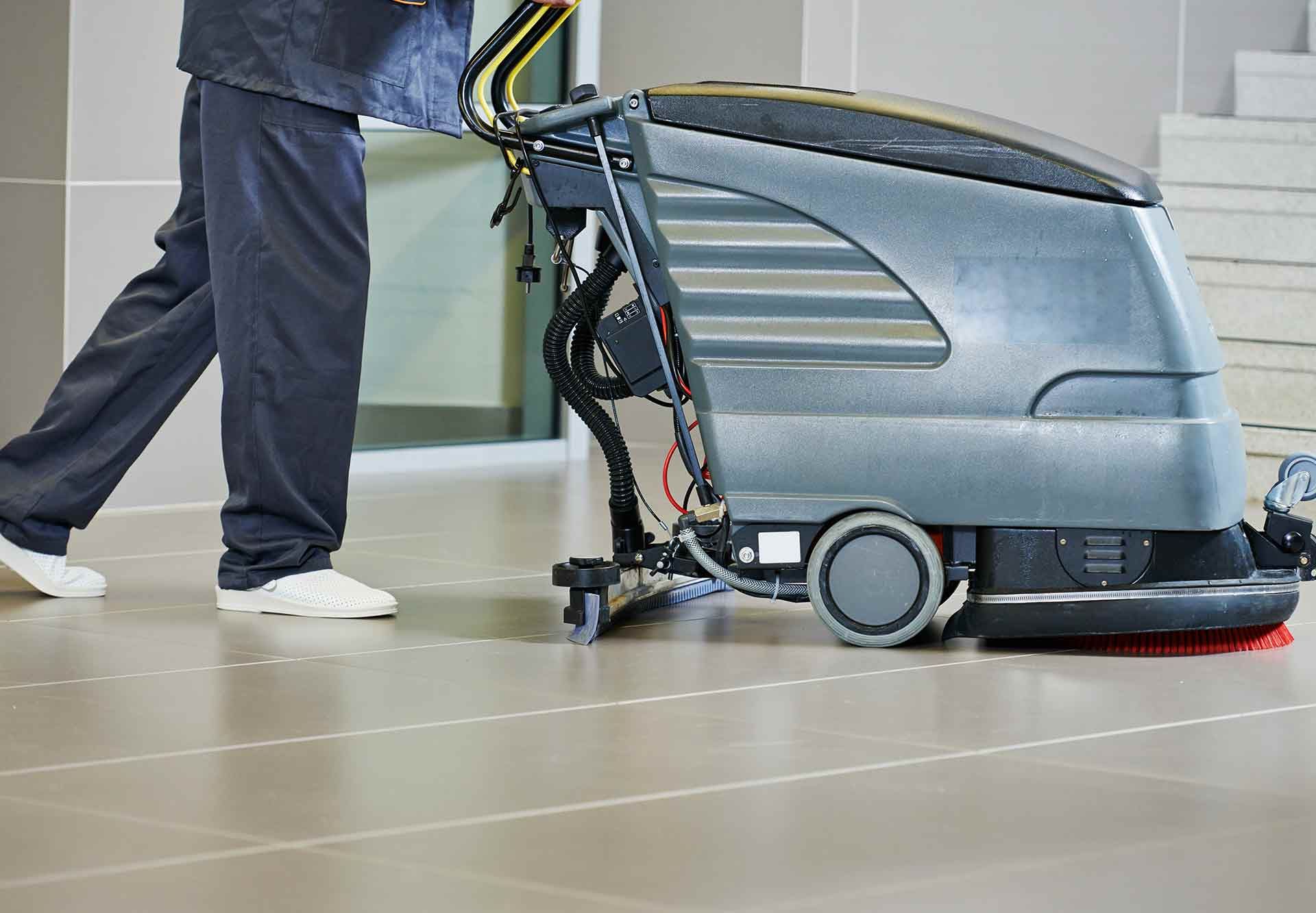 Une personne pousse une machine de nettoyage de sol grise sur un sol carrelé dans un couloir d'immeuble.
