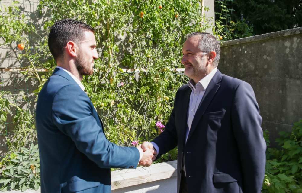 Deux hommes en costume se serrent la main dans un jardin ensoleillé.