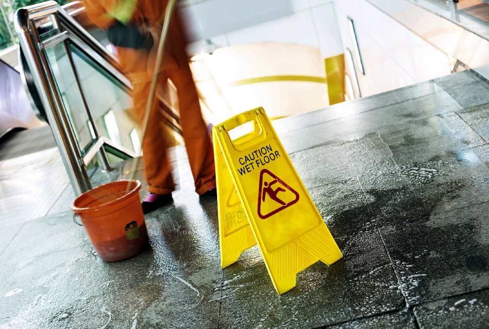 Panneau jaune « sol mouillé » et agent d'entretien passant la serpillière sur un sol intérieur brillant près d'un seau et d'un escalier.