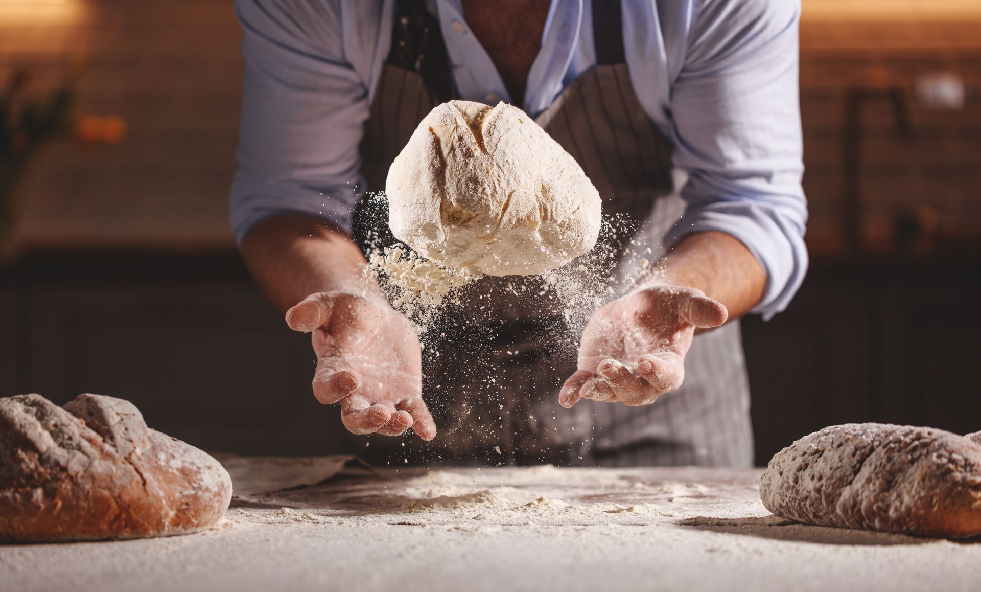 Le boulanger fait tournoyer la pâte en l'air, la table est saupoudrée de farine.