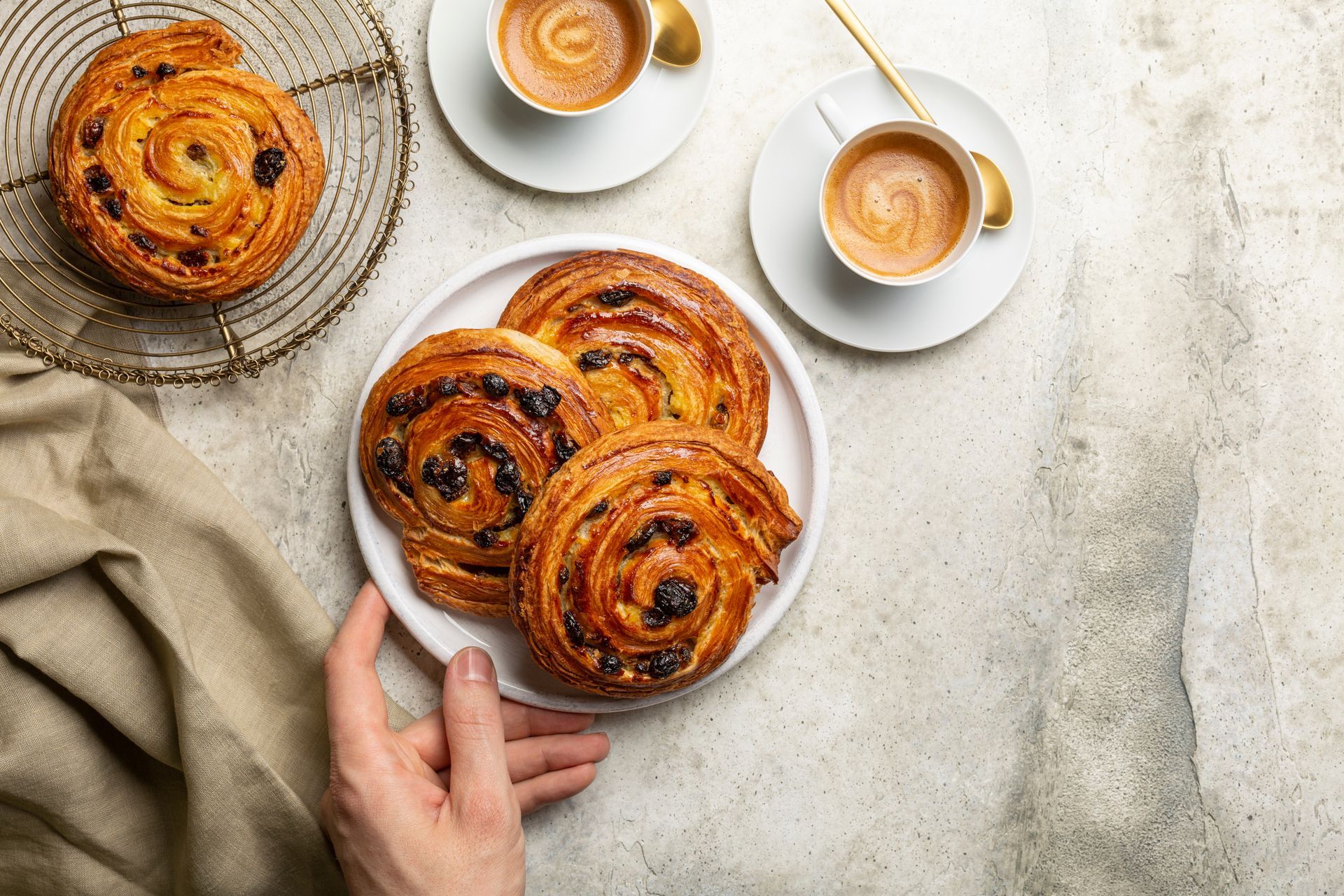 Une personne tient une assiette avec trois pâtisseries tourbillonnées aux raisins secs, deux tasses à café et une autre pâtisserie sur une grille.