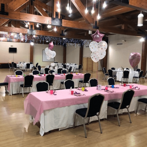 Salón de eventos con mesas preparadas para fiesta, decorado con manteles rosas, globos y serpentinas.