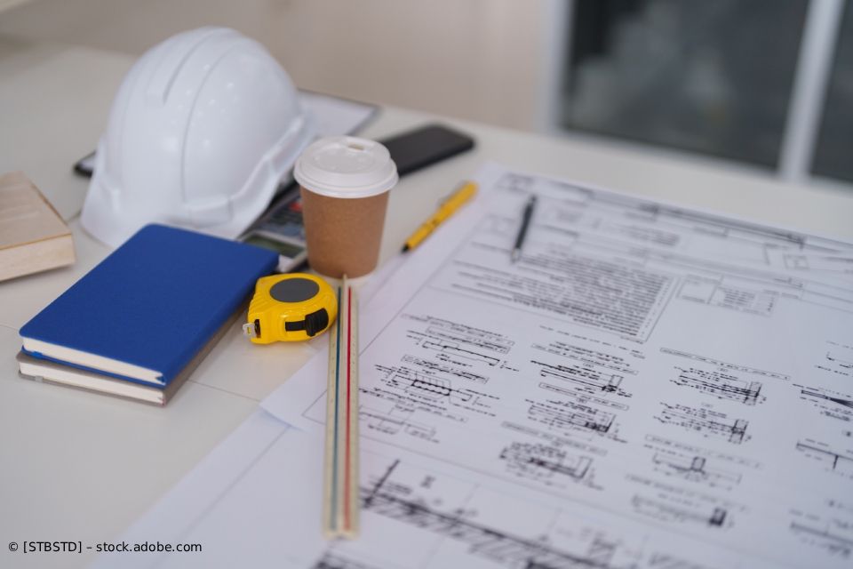 Baupläne, Schutzhelm, Kaffee und Werkzeug auf einem Tisch.