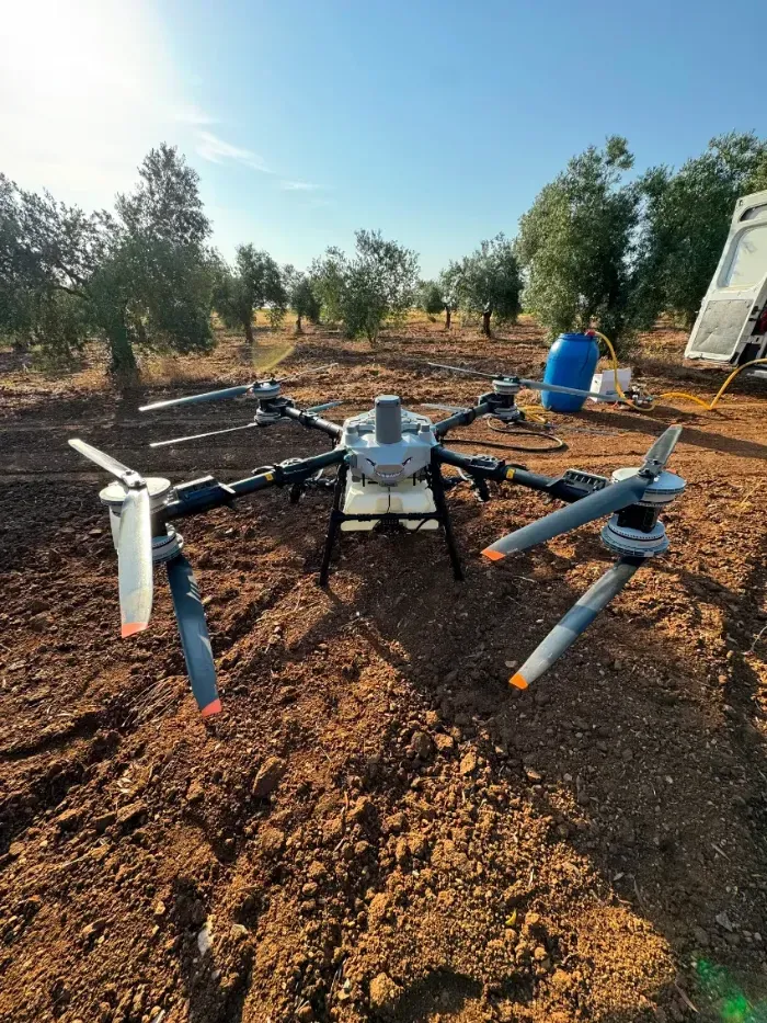 Gran dron sobre un campo seco de tierra roja con árboles y un barril azul al fondo.