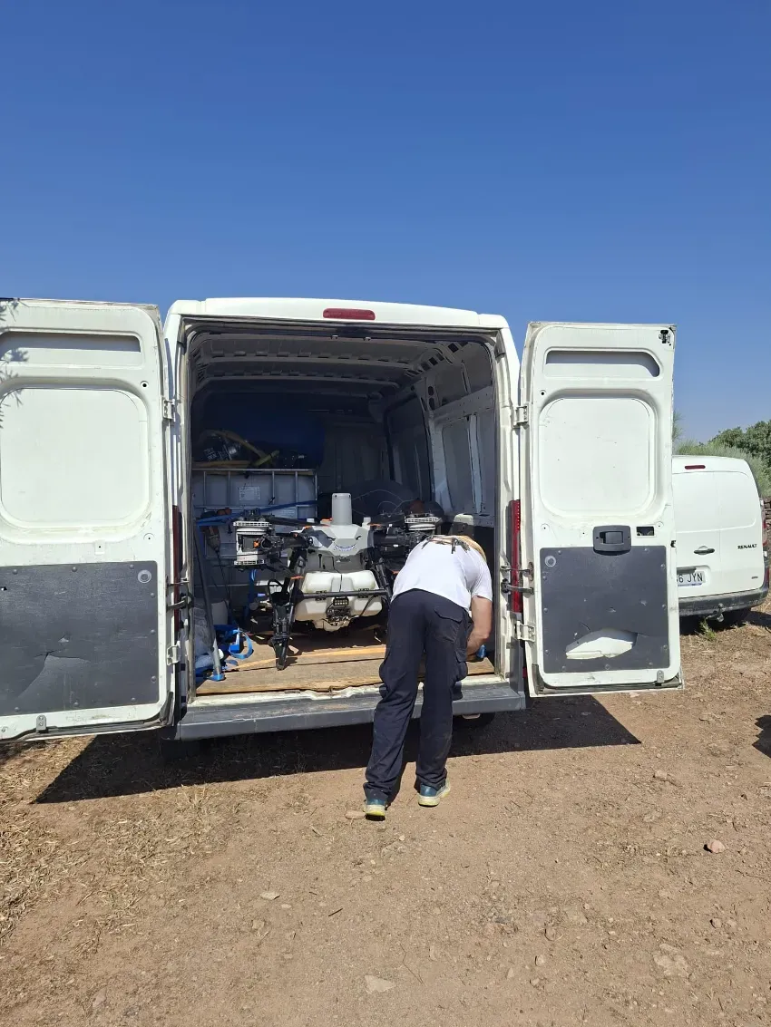 Una persona carga equipo en una furgoneta blanca con las puertas traseras abiertas en un terreno baldío.