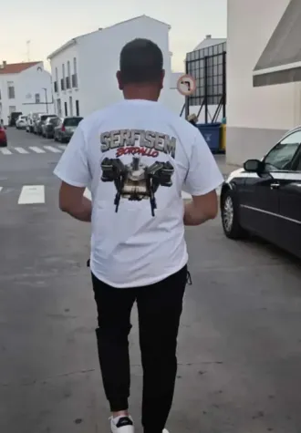 Una persona se aleja caminando por una calle de la ciudad, vistiendo una camiseta blanca estampada y pantalones negros.