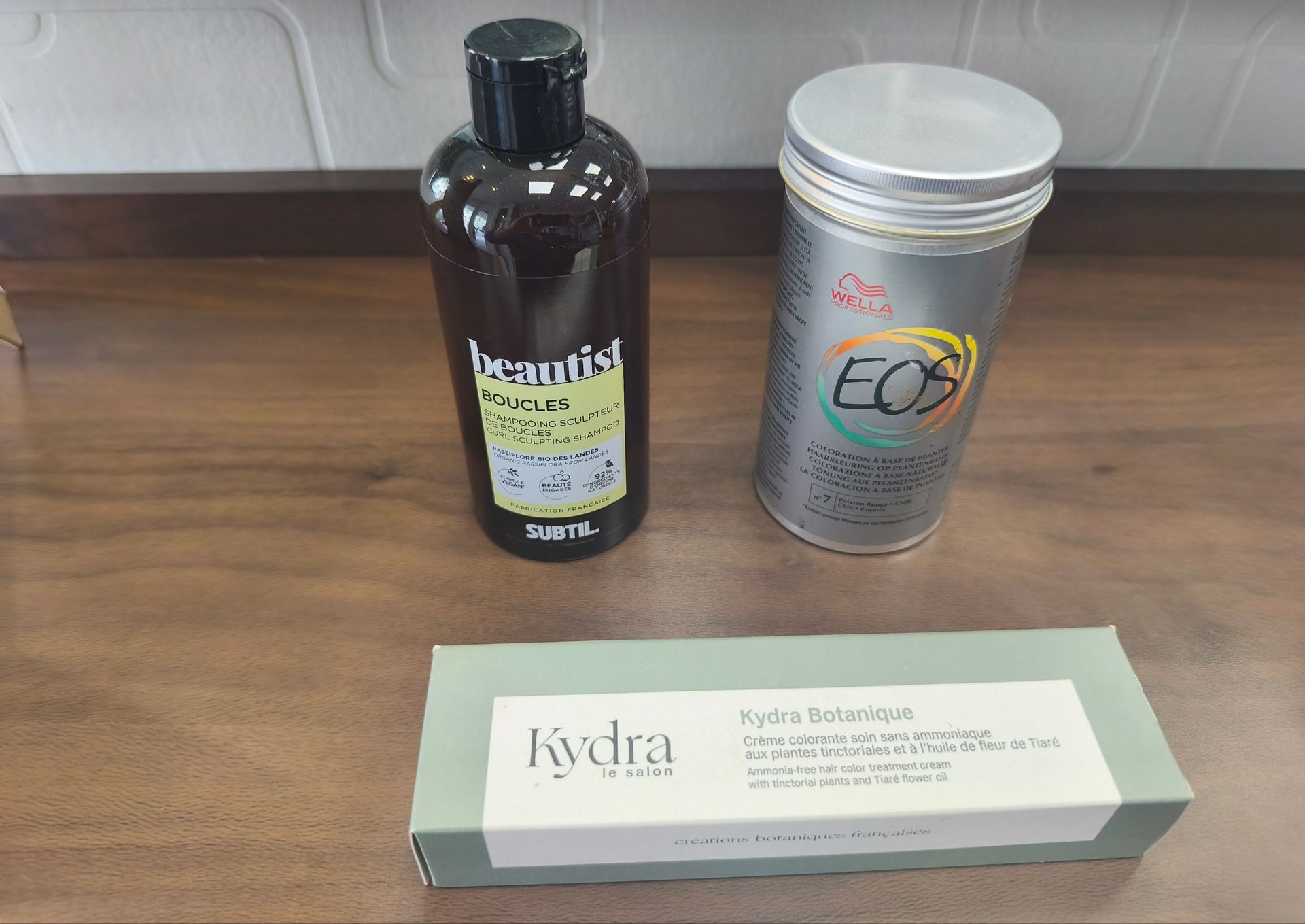 Trois emballages de produits sur une table en bois : une bouteille noire, un flacon argenté et une boîte blanche.