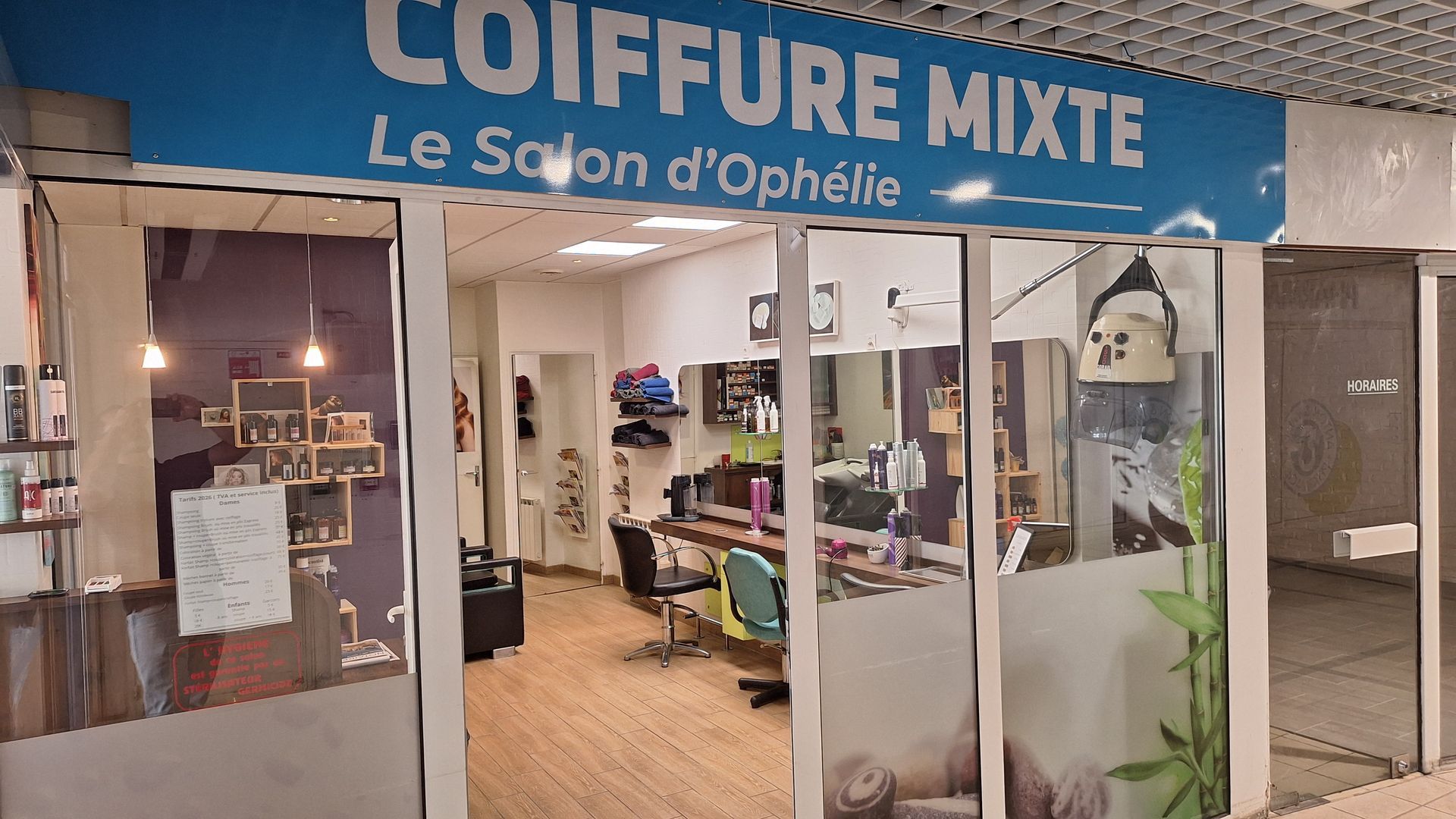Façade de salon de coiffure avec enseigne bleue, entrée vitrée et fauteuils à l'intérieur.