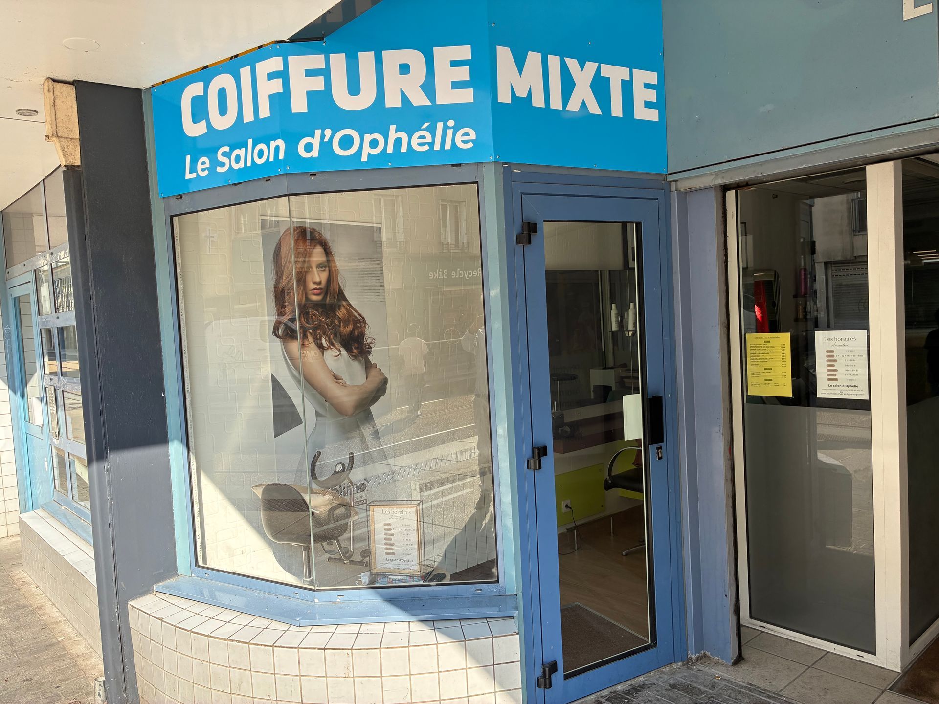 Devanture bleue du salon de coiffure Coiffure Mixte, avec une affiche représentant une femme dans la vitrine et l’entrée ouverte