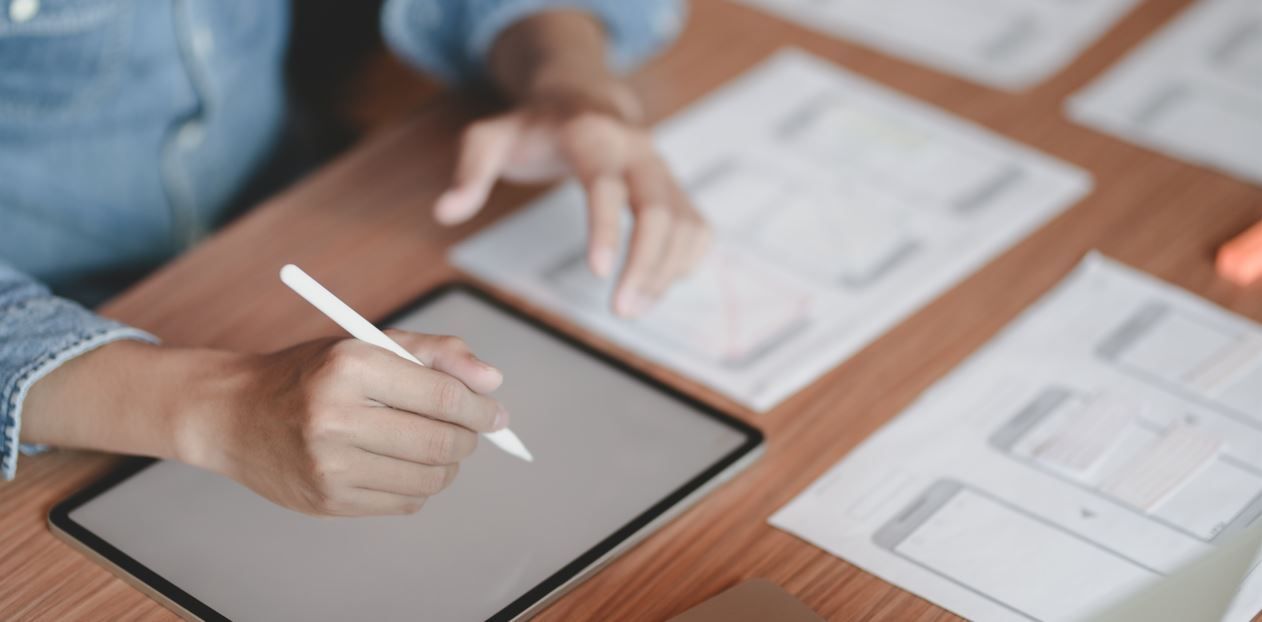 Persona dibujando en una tableta con un lápiz, rodeada de papeles con diagramas sobre una mesa de madera.