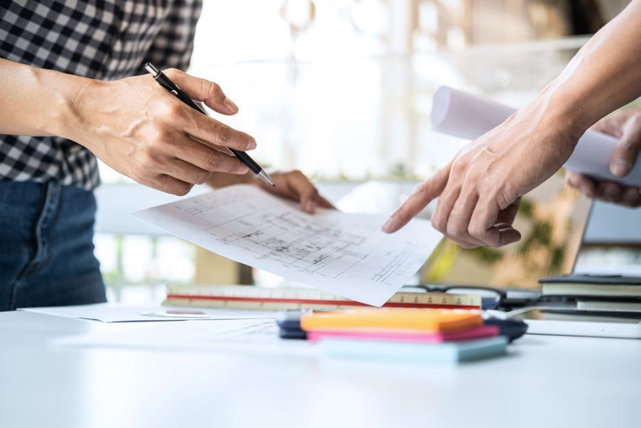 Dos personas revisando planos arquitectónicos en un escritorio, una señalando y otra sosteniendo un bolígrafo.